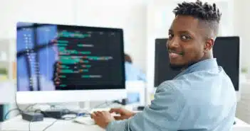 Happy guy looking at you while sitting by workplace and working with software