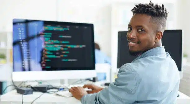 Happy guy looking at you while sitting by workplace and working with software